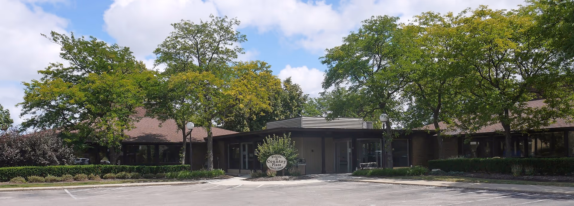 Exterior view of Wickliffe Country Place, a single-story building surrounded by trees and greenery under a partly cloudy sky, with a parking lot in front.