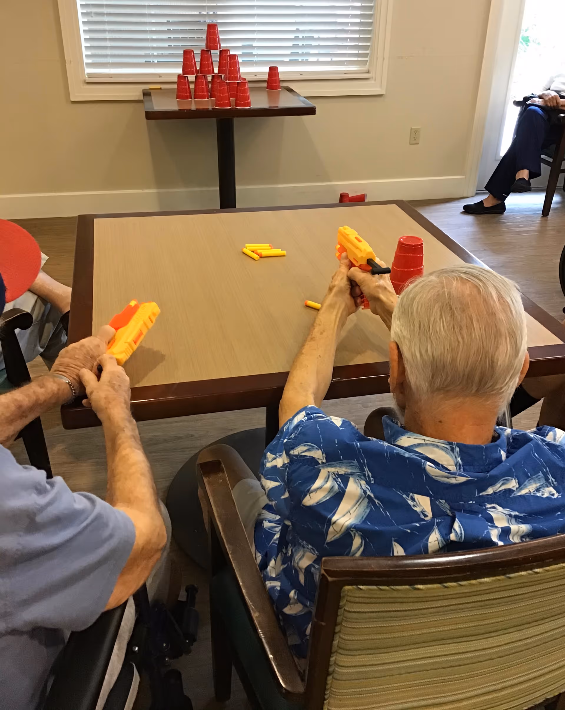 Two elderly men sitting at a table in a room, aiming toy dart guns at a stack of red plastic cups arranged on a smaller table in front of a window with blinds. One man is wearing a blue patterned shirt and the other a light blue shirt and red cap. Another person is partially visible sitting in the background near a door.