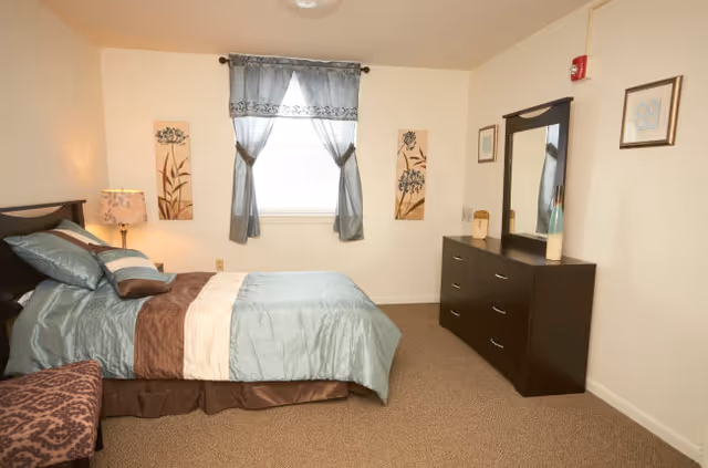 A neatly made bed with blue and brown bedding in a bedroom. There is a window with blue curtains above the bed's headboard. On the right side of the room, there is a dark wooden dresser with a mirror and decorative items on top. The walls are light-colored and decorated with framed artwork.