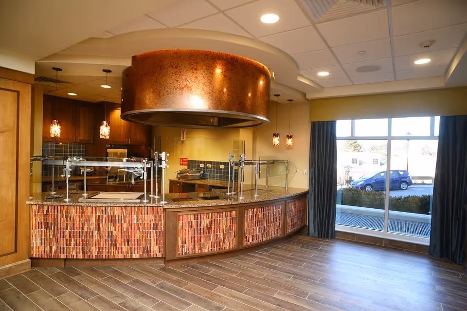 Interior view of a kitchen area in a senior living facility with a curved counter featuring a mosaic tile front and granite countertop. Above the counter is a large copper-colored vent hood. The kitchen has wooden cabinets, pendant lights, and a large window with dark curtains letting in natural light.