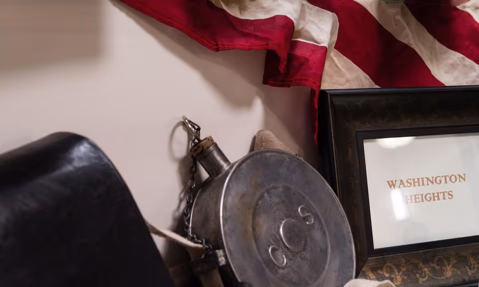 Close-up view of a vintage metal canteen with the letters 'C O S' embossed on it, resting against a wall with a partially visible American flag draped above and a framed sign that reads 'WASHINGTON HEIGHTS'.