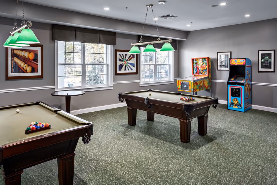 A game room with two pool tables, each with billiard balls arranged on the felt surface. The room has green carpet, gray walls, and two large windows with brown valances. There are three green hanging lights above each pool table. In the corner, there is a colorful pinball machine and a classic Pac-Man arcade game. Framed pictures hang on the walls.
