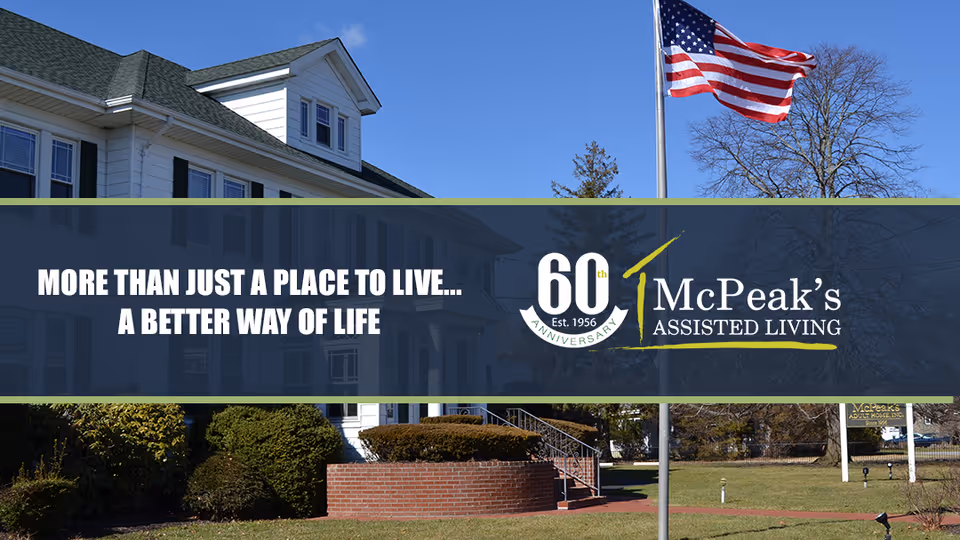 Exterior view of McPeak's Assisted Living facility showing a white building with multiple windows, a green roof, an American flag on a flagpole, and landscaped bushes and trees under a clear blue sky.