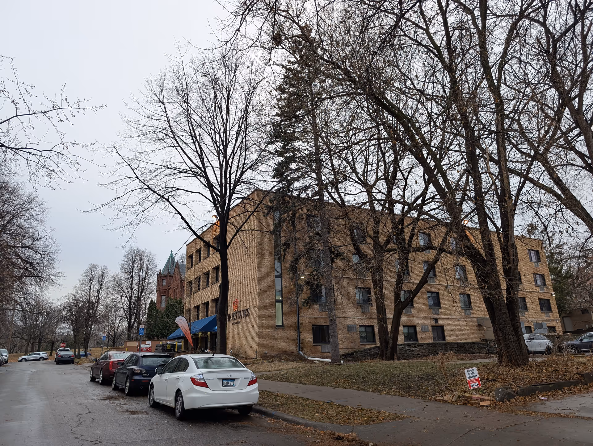 Exterior view of a four-story brick building named The Estates at Chateau, with several parked cars along the street and leafless trees surrounding the area on a cloudy day.