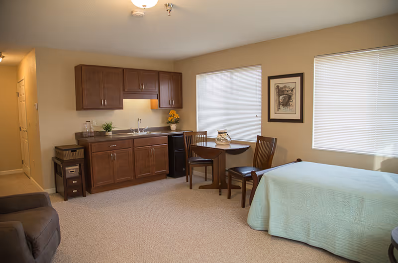 A cozy senior living studio apartment featuring a small kitchenette with wooden cabinets, a sink, and a mini refrigerator. There is a round wooden dining table with two chairs near two windows with blinds. A bed with a light blue quilt is positioned near the windows, and a comfortable armchair is visible in the foreground. The walls are painted beige, and there is a framed picture hanging between the windows.