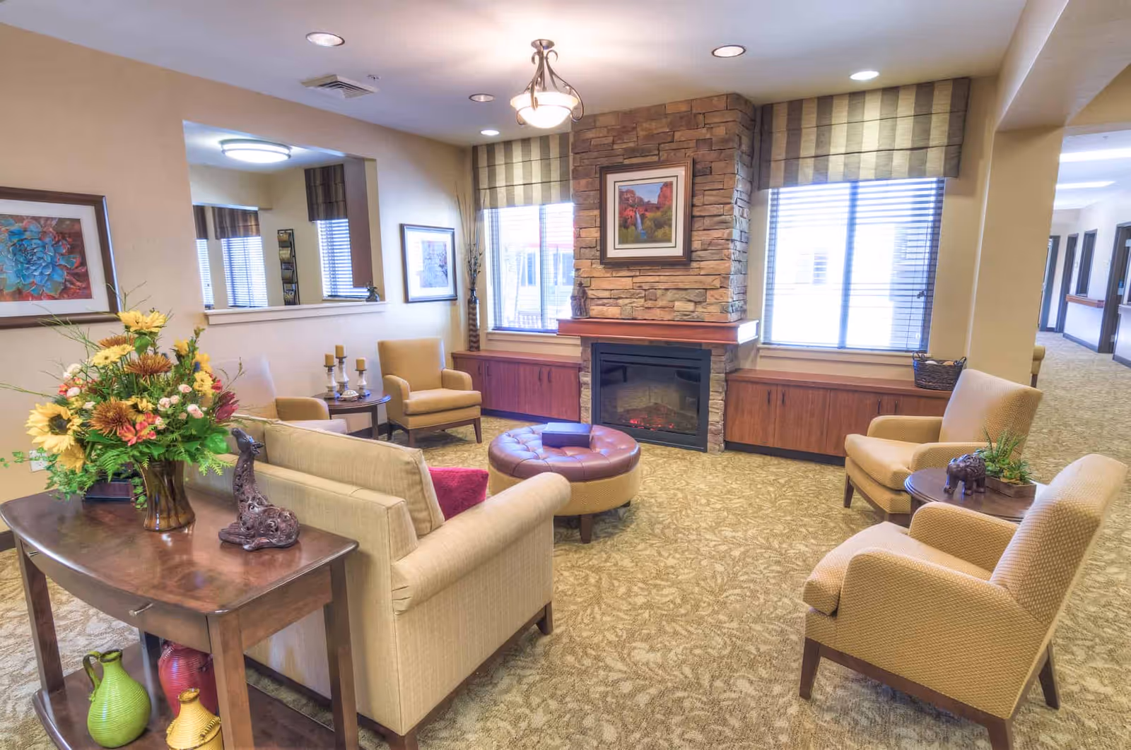 A cozy living room area in a senior living facility with beige upholstered chairs and a sofa arranged around a round ottoman. There is a stone fireplace with a framed picture above it, two windows with striped valances, and a wooden console table with a large floral arrangement and decorative items. The room has warm lighting and patterned carpet.