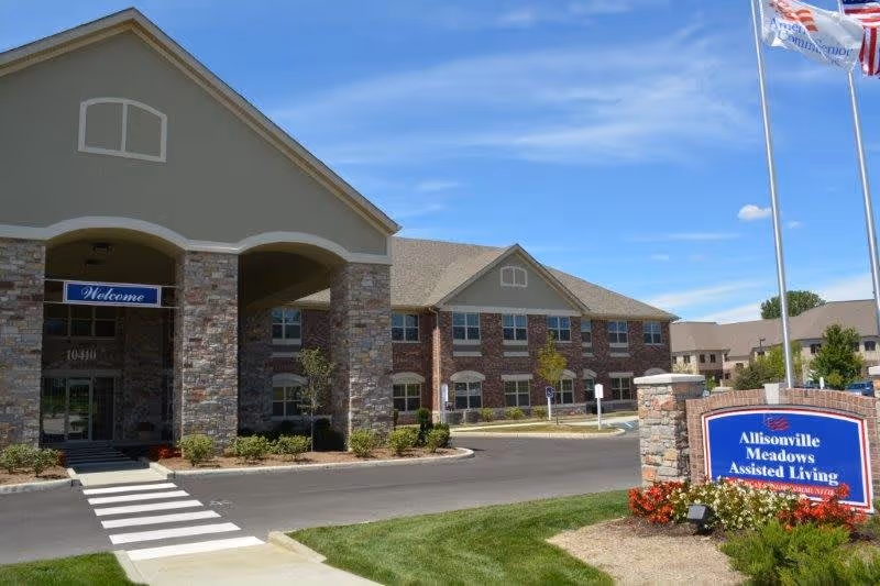 Exterior view of Allisonville Meadows Assisted Living facility showing a two-story building with stone and brick facade, a covered entrance with a 'Welcome' sign, a driveway, and a landscaped area with a sign displaying the facility's name. Three flagpoles with flags are visible on the right side under a clear blue sky.