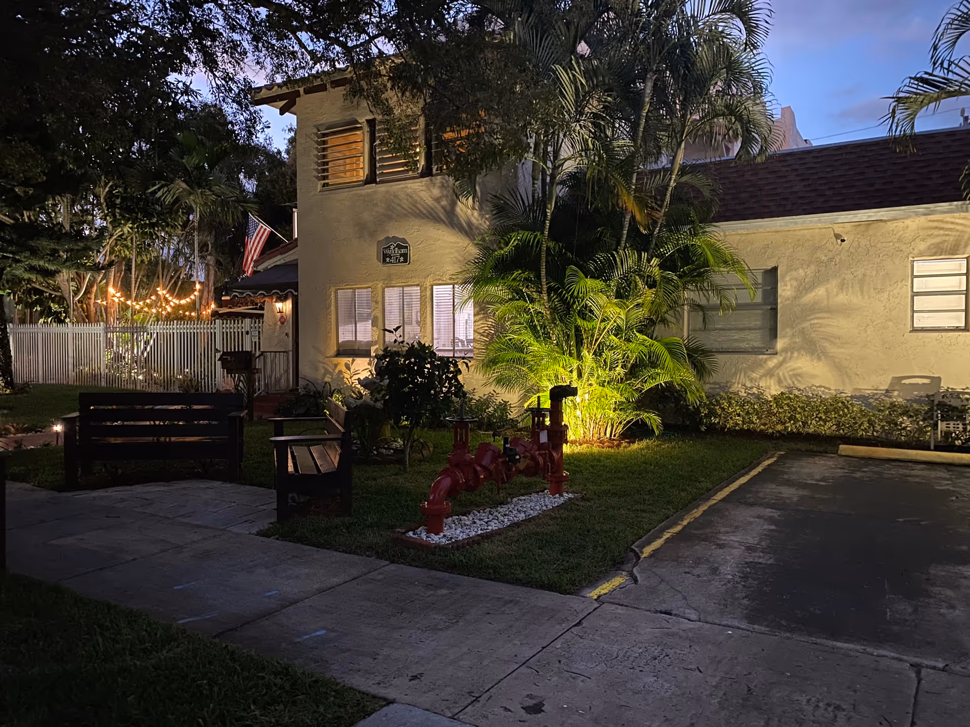 Evening view of the exterior of a building at Wyndham House with illuminated palm trees, benches, a small garden area, and a parking space. The building has a beige facade with windows and an American flag near the entrance.