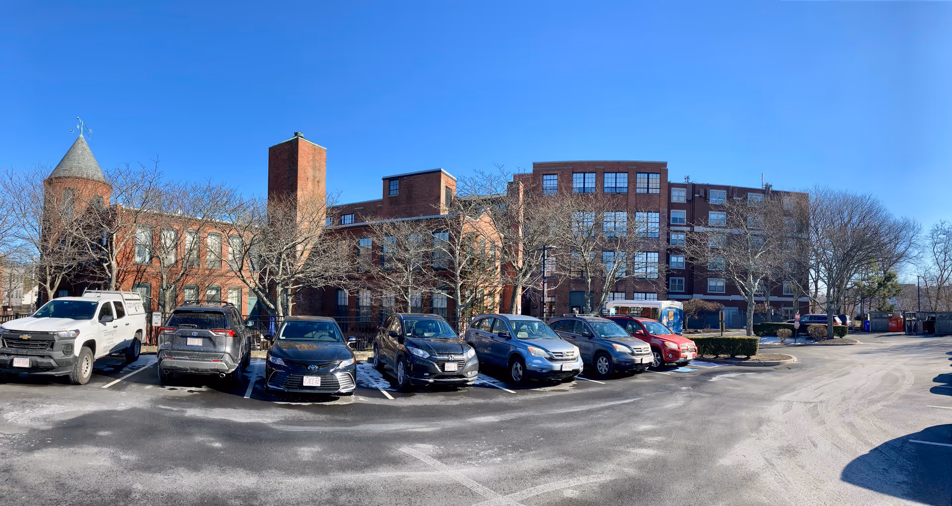 Wide exterior view of a multi-story brick building with large windows, surrounded by leafless trees and a parking lot with several parked cars under a clear blue sky.