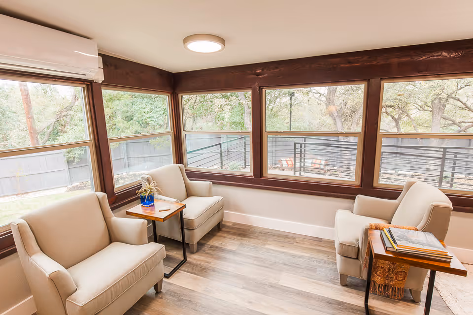 A bright sunroom with large windows on two sides showing a view of trees and a fenced yard. The room has three beige armchairs arranged around two small wooden side tables, one with a small plant and the other with books. The floor is wood laminate and there is a white ceiling light and an air conditioning unit above the windows.