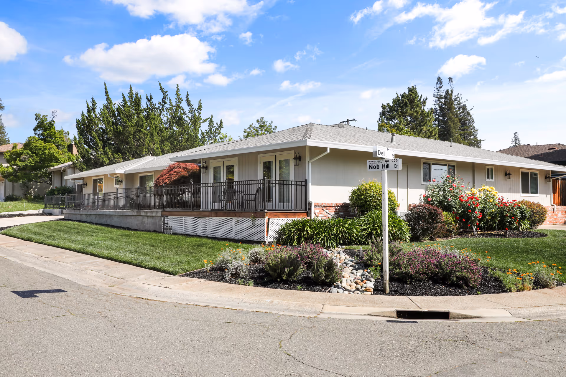 Exterior view of a single-story care home building with a gray roof and light-colored walls. The building has a porch with black railings and outdoor chairs. The surrounding area features well-maintained green lawns, flower beds with colorful flowers, and various shrubs. A street sign at the corner reads 'Dell' and 'Nob Hill Dr'. The sky is blue with scattered clouds.
