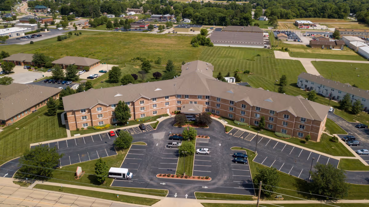 Aerial view of Cambridge House of Swansea, a large three-story brick senior living facility with a U-shaped layout, surrounded by green lawns and parking lots with several cars parked. The building is situated in a suburban area with other buildings and trees in the background.
