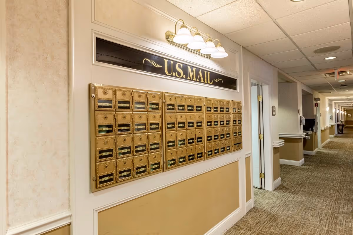 A hallway in a senior living facility with a wall-mounted set of gold-colored U.S. Mailboxes labeled 'U.S. MAIL' above. The hallway has beige walls with white trim, carpeted flooring, and ceiling lights. Doors and other hallway features are visible further down the corridor.