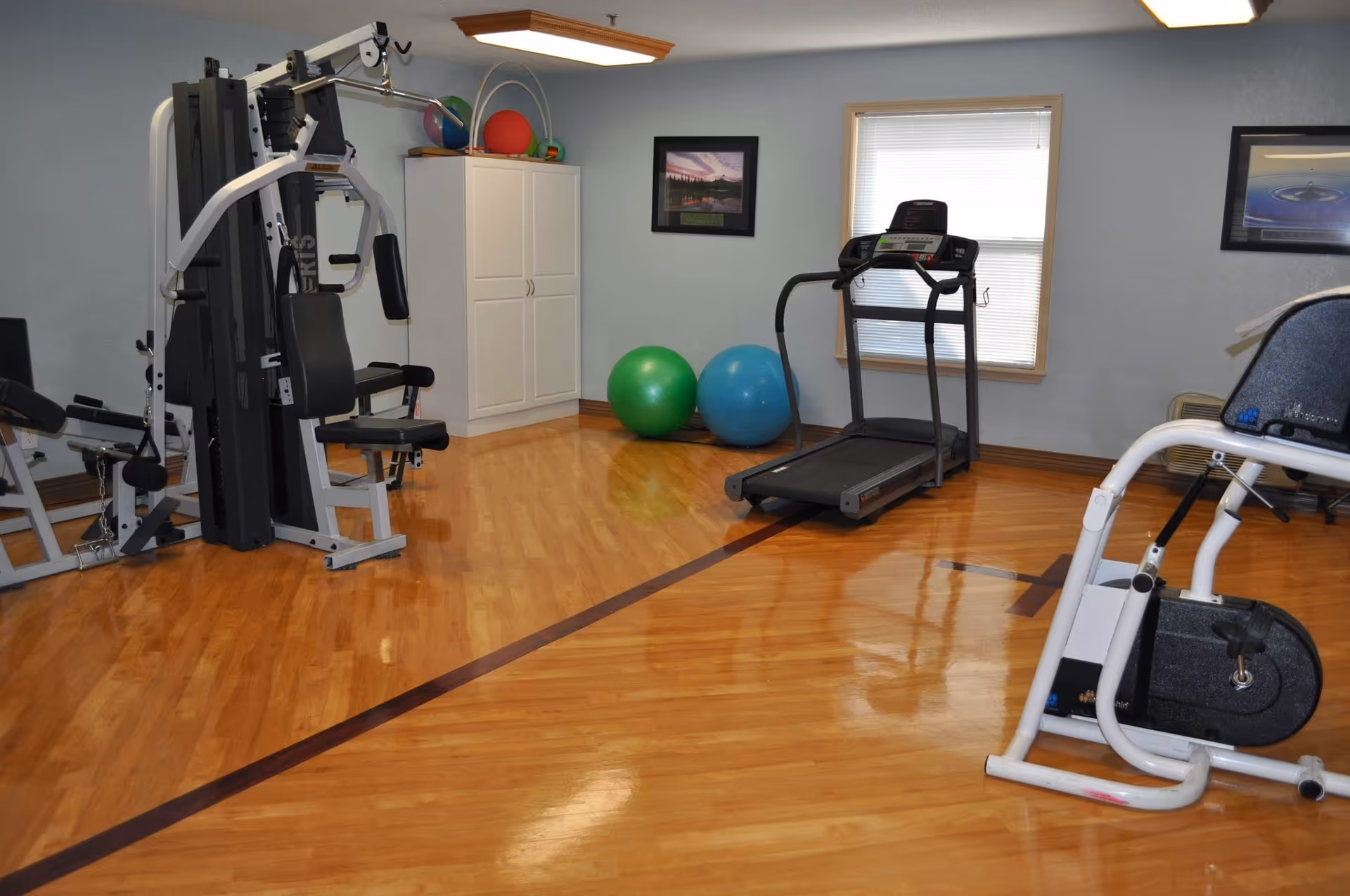 A fitness room with exercise equipment including a multi-station gym machine, a treadmill, an exercise bike, and two large exercise balls. The room has polished wooden floors, light blue walls, a window with blinds, and a white cabinet in the corner.