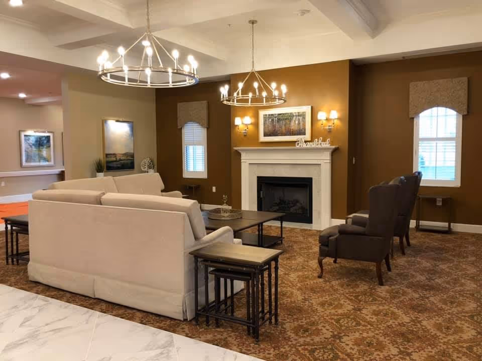 A cozy senior living facility common area with beige sofas arranged around a dark wooden coffee table, two dark armchairs, a white fireplace with a framed picture above it, and two large circular chandeliers hanging from the ceiling. The room has warm brown walls, patterned carpet, and windows with beige valances.