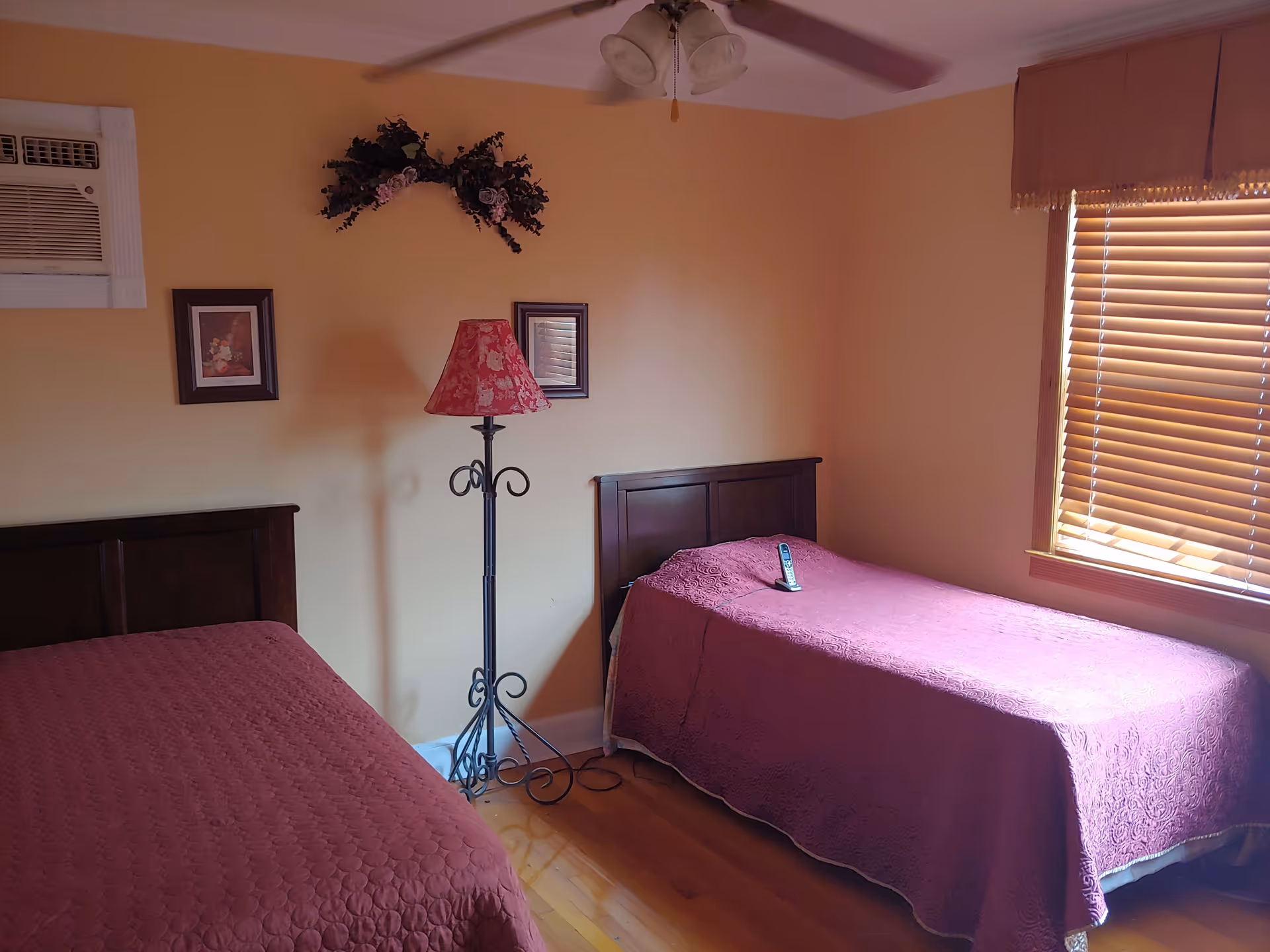 A bedroom with two single beds covered in matching purple bedspreads. Between the beds is a tall floor lamp with a red floral lampshade. Above the lamp on the wall is a decorative floral arrangement and two framed pictures. There is a window with wooden blinds on the right side, and an air conditioning unit is installed on the left wall.