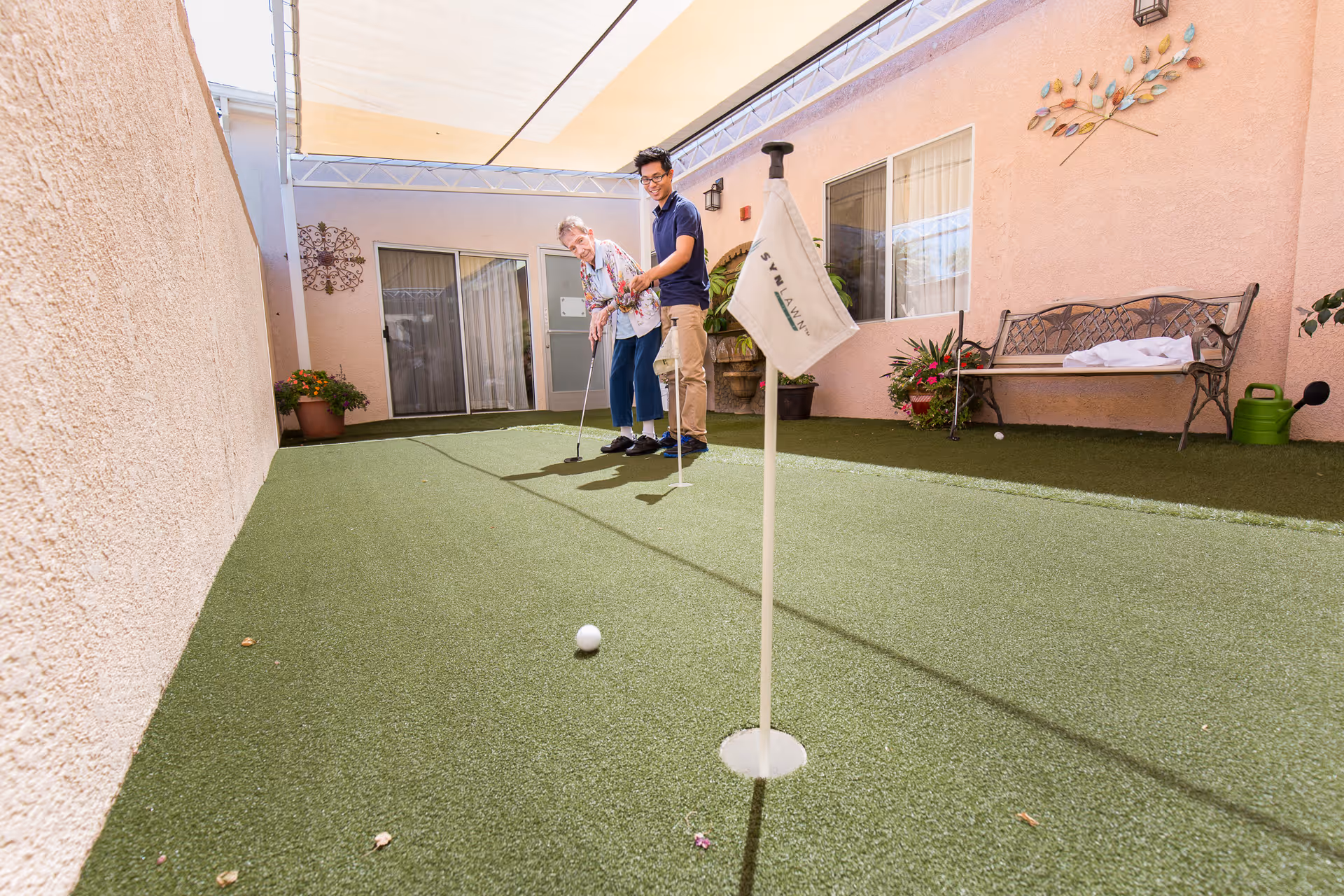 An elderly woman and a younger man are playing mini golf on a small artificial turf putting green in an outdoor courtyard area of a healthcare facility. The courtyard is surrounded by light pink walls with windows and doors, decorated with potted plants, a bench, and wall art. A white flag with the word 'SYLVAN' is visible near the hole on the putting green.