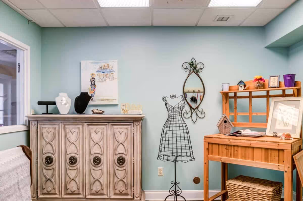 A cozy interior room with light blue walls featuring a decorative wooden cabinet with jewelry displays on top, a wire dress form mannequin, an ornate wall mirror, and a wooden desk with shelves holding small decorative items and a wicker basket underneath.