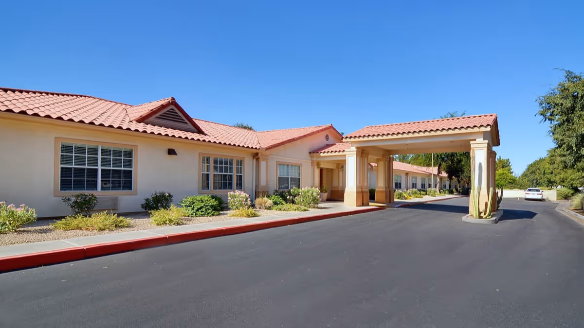 Exterior view of a single-story building with a red tile roof and beige walls, featuring multiple windows and a covered entrance supported by columns. The building is surrounded by landscaped bushes and plants, with a clear blue sky overhead and a paved driveway in front.