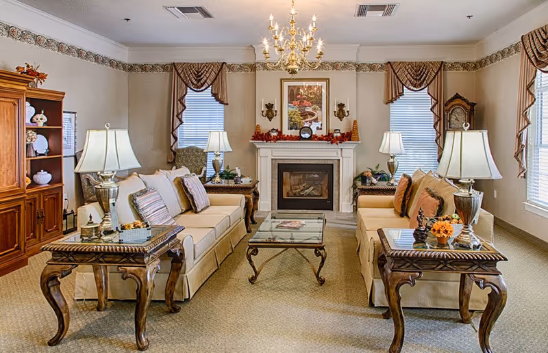 A cozy senior living facility common area with two beige sofas facing each other, each adorned with multiple cushions. Between the sofas is a glass coffee table with ornate wooden legs. On either side of the sofas are matching wooden side tables with decorative lamps and small floral arrangements. The room features a white fireplace with autumn-themed decorations on the mantel, a framed picture above it, and two windows with brown valances. A chandelier hangs from the ceiling, and a wooden cabinet with decorative items is visible on the left side.