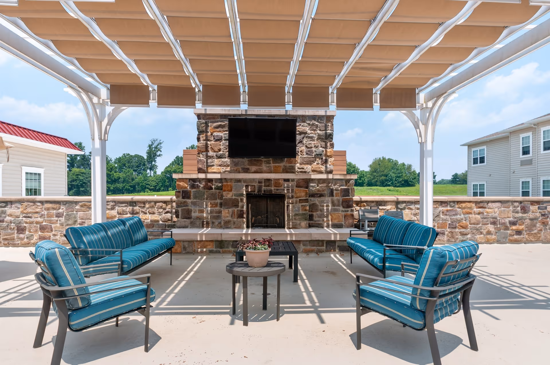 Outdoor seating area with blue cushioned chairs and sofas arranged around a stone fireplace with a mounted flat-screen TV under a pergola with beige fabric shades. Stone walls and greenery are visible in the background.