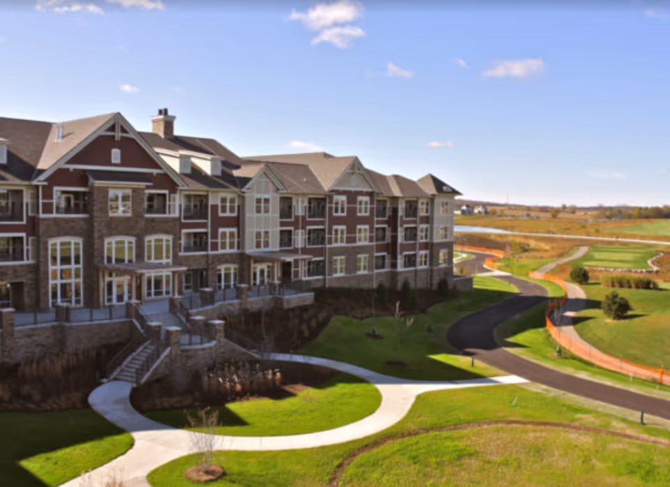 Exterior view of a large multi-story senior living facility building with stone and siding facade, surrounded by green lawns, curved sidewalks, and a paved road under a clear blue sky.