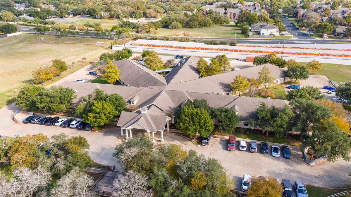Aerial view of West Oaks Nursing and Rehabilitation Center showing a large, single-story building with a central entrance and multiple wings surrounded by trees and parking lots with several cars. The facility is situated near a residential area and a storage unit complex.
