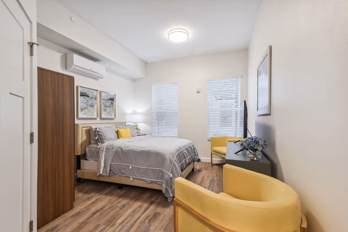 A bright and modern bedroom with a queen-sized bed dressed in gray and white bedding, two yellow armchairs, a dark wooden dresser with a TV on top, two windows with white blinds, and two framed landscape paintings on the wall above the bed. The room has wood flooring and white walls.