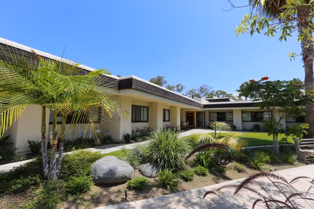 Exterior view of a single-story building with a flat roof surrounded by landscaped greenery including palm trees, bushes, and rocks under a clear blue sky.