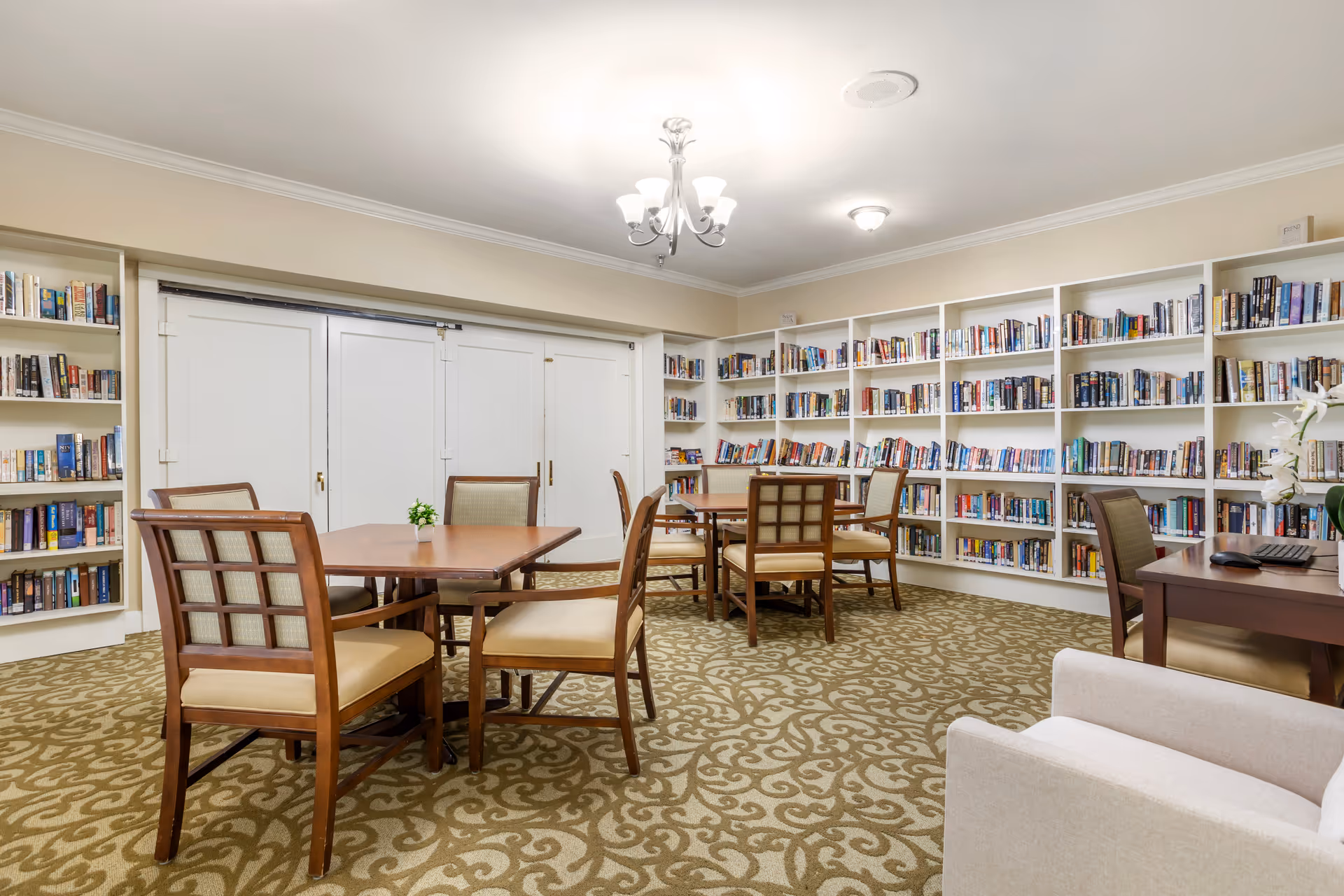 A cozy library room with multiple wooden tables and cushioned chairs arranged on a patterned carpet. The walls are lined with white bookshelves filled with books. The room is well-lit with ceiling lights and has a calm, inviting atmosphere.