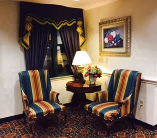 A cozy interior corner featuring two striped upholstered armchairs with wooden frames, a small round wooden table between them holding a lamp and a floral arrangement. The wall behind has a framed painting of flowers and a window with dark curtains and decorative valance.