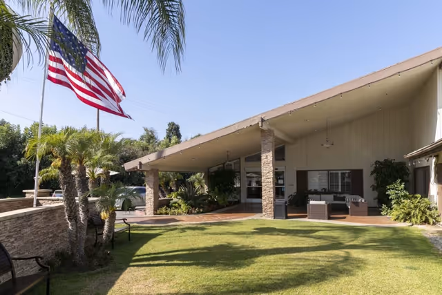 Exterior view of St. Elizabeth Healthcare & Rehab building with a slanted roof supported by stone pillars, a well-maintained lawn, palm trees, and an American flag flying on a flagpole.