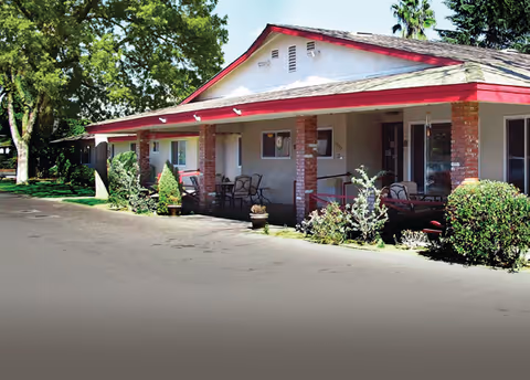Exterior view of a single-story building with a red-trimmed roof and a covered porch area featuring outdoor seating and potted plants, surrounded by trees and greenery.