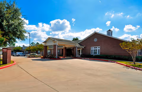 Exterior view of a single-story brick building with a covered entrance and a driveway in front. The sky is blue with scattered clouds, and there are trees and landscaping around the building.