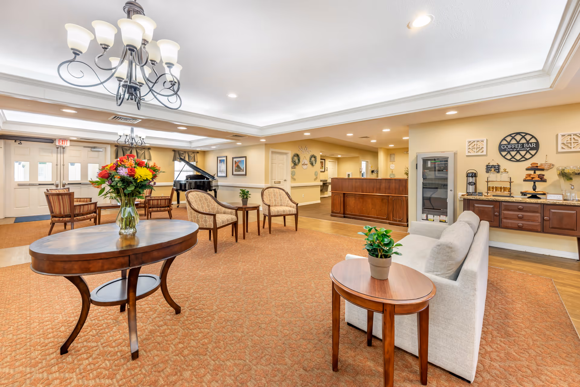 A spacious and well-lit senior living facility common area with beige walls and carpeted floor. The room features a round wooden table with a vase of colorful flowers, several upholstered chairs, a white sofa with a small wooden side table holding a potted plant, and a grand piano in the background. There is a coffee bar area with a coffee machine, pastries, and cups on a wooden counter. The ceiling has recessed lighting and a decorative chandelier.
