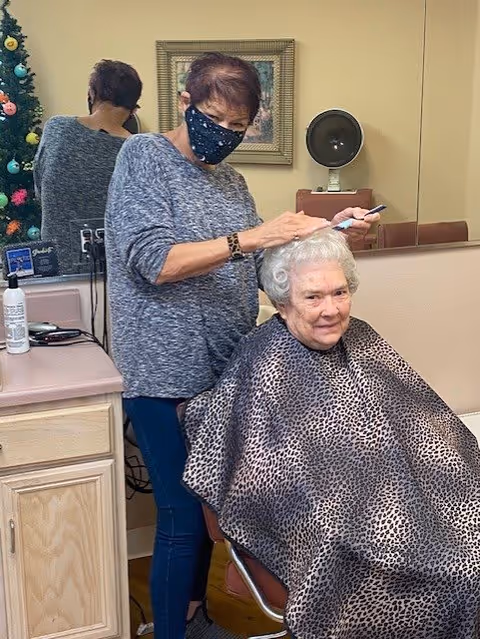 An elderly woman with white hair sits in a salon chair wearing a leopard print cape while a hairstylist wearing a face mask styles her hair. The setting appears to be a salon area with a mirror, countertop, and hair care products visible.