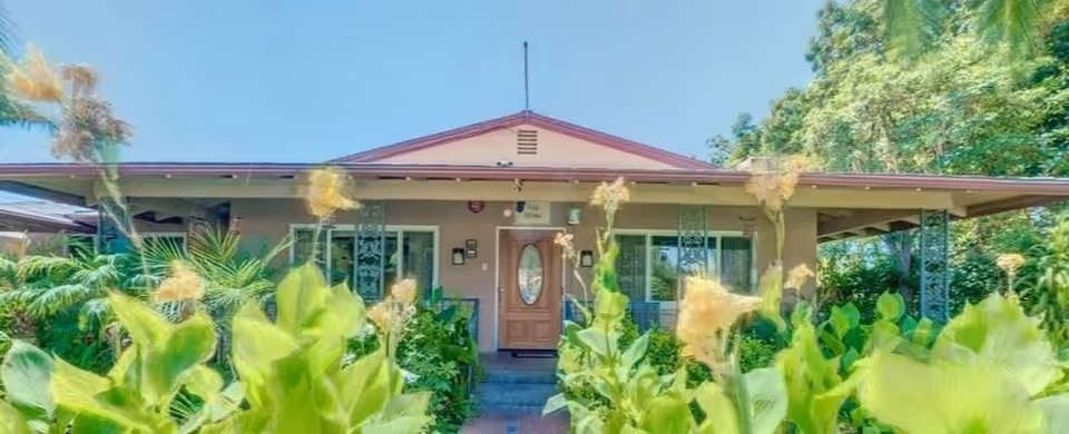 Front exterior view of a single-story building with a peaked roof, a wooden door with an oval glass window, and surrounded by lush green plants and trees under a clear blue sky.