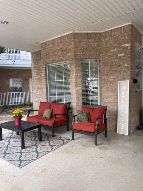 Outdoor seating area with two red cushioned chairs and a matching loveseat around a dark wooden coffee table with a potted yellow flower on top. The seating is placed on a patterned outdoor rug in front of a brick wall with two windows. A tall vertical sign next to the seating reads 'WELCOME'.