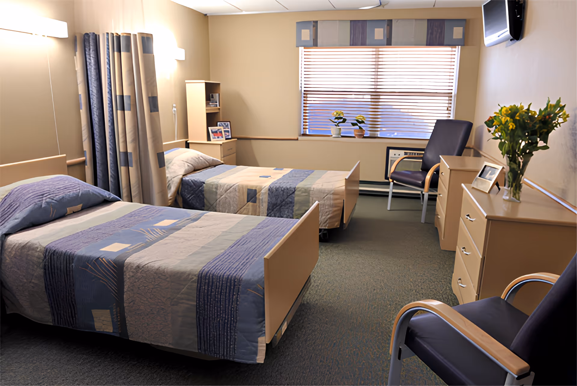 A well-lit room with two single beds separated by a curtain, each bed covered with blue and beige patterned bedspreads. The room has beige walls, a window with blinds, two chairs with wooden armrests, a dresser with a vase of yellow flowers, and a small cabinet with framed photos. A wall-mounted TV is visible in the corner.