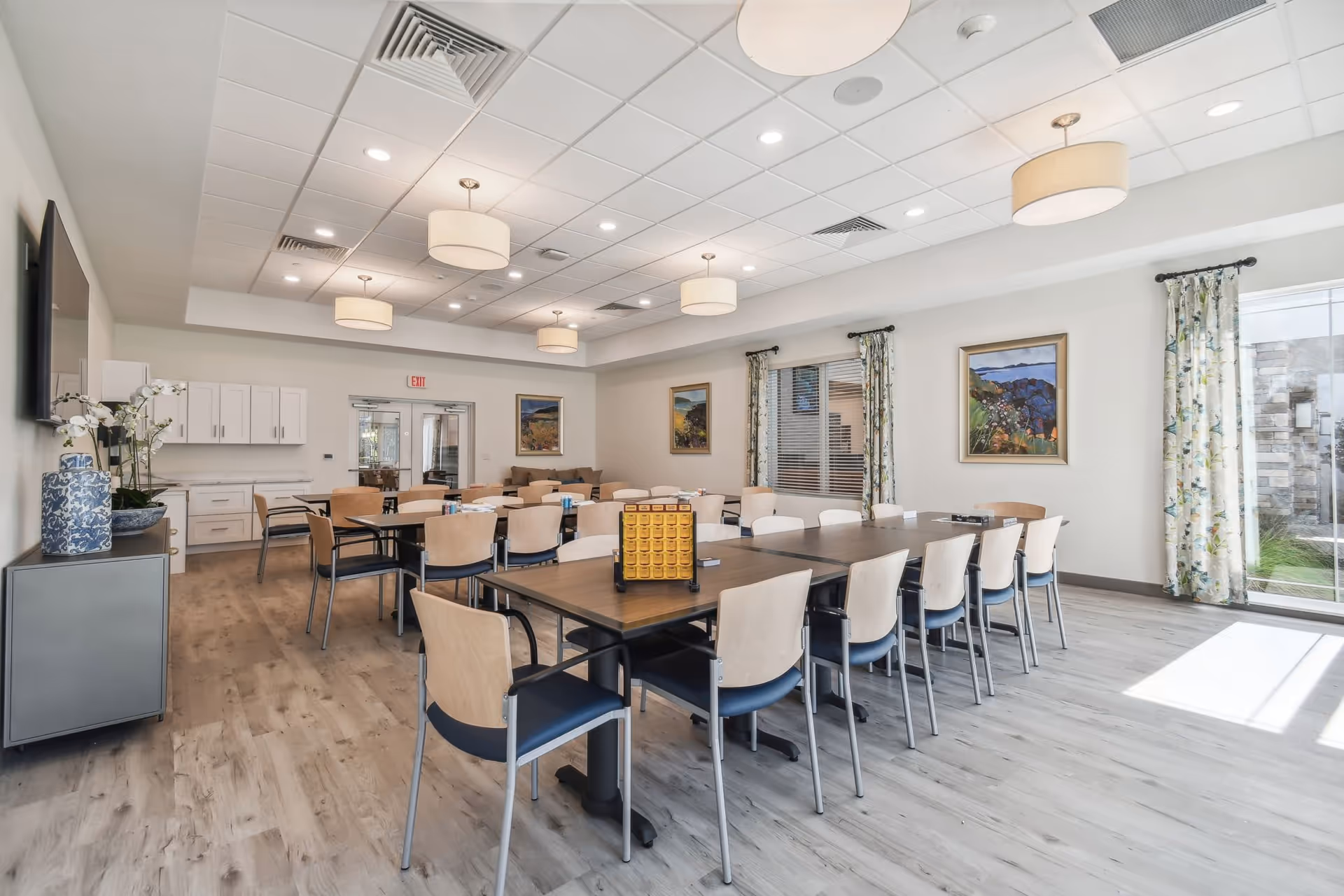A bright and spacious dining room with multiple wooden tables and chairs arranged neatly. The room features light-colored wood flooring, white walls, and a ceiling with recessed lighting and hanging pendant lamps. There are framed landscape paintings on the walls and large windows with floral curtains allowing natural light to enter. A cabinet with decorative vases and a flat-screen TV is visible on the left side.