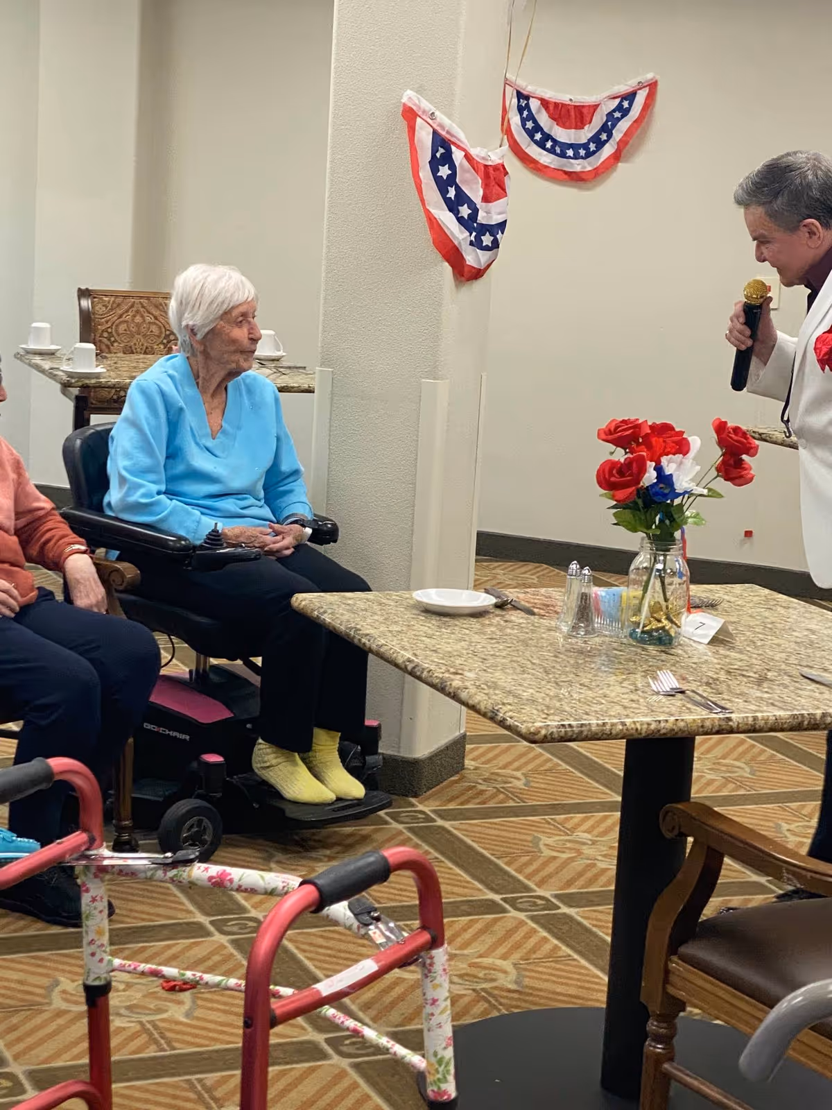 An elderly woman in a blue sweater sitting in a motorized wheelchair in a common area decorated with red, white, and blue bunting. A man in a white jacket is speaking into a microphone near a table with a vase of red, white, and blue flowers. A walker is visible in the foreground.