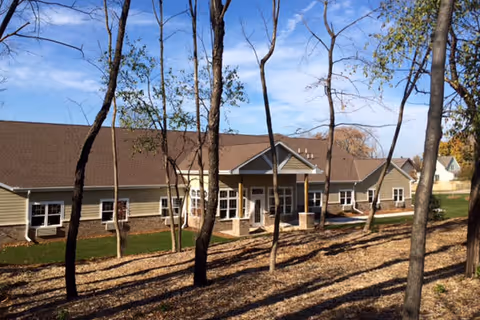 Exterior view of a single-story building with a brown roof and beige siding, surrounded by leafless trees and a grassy area under a blue sky with some clouds.