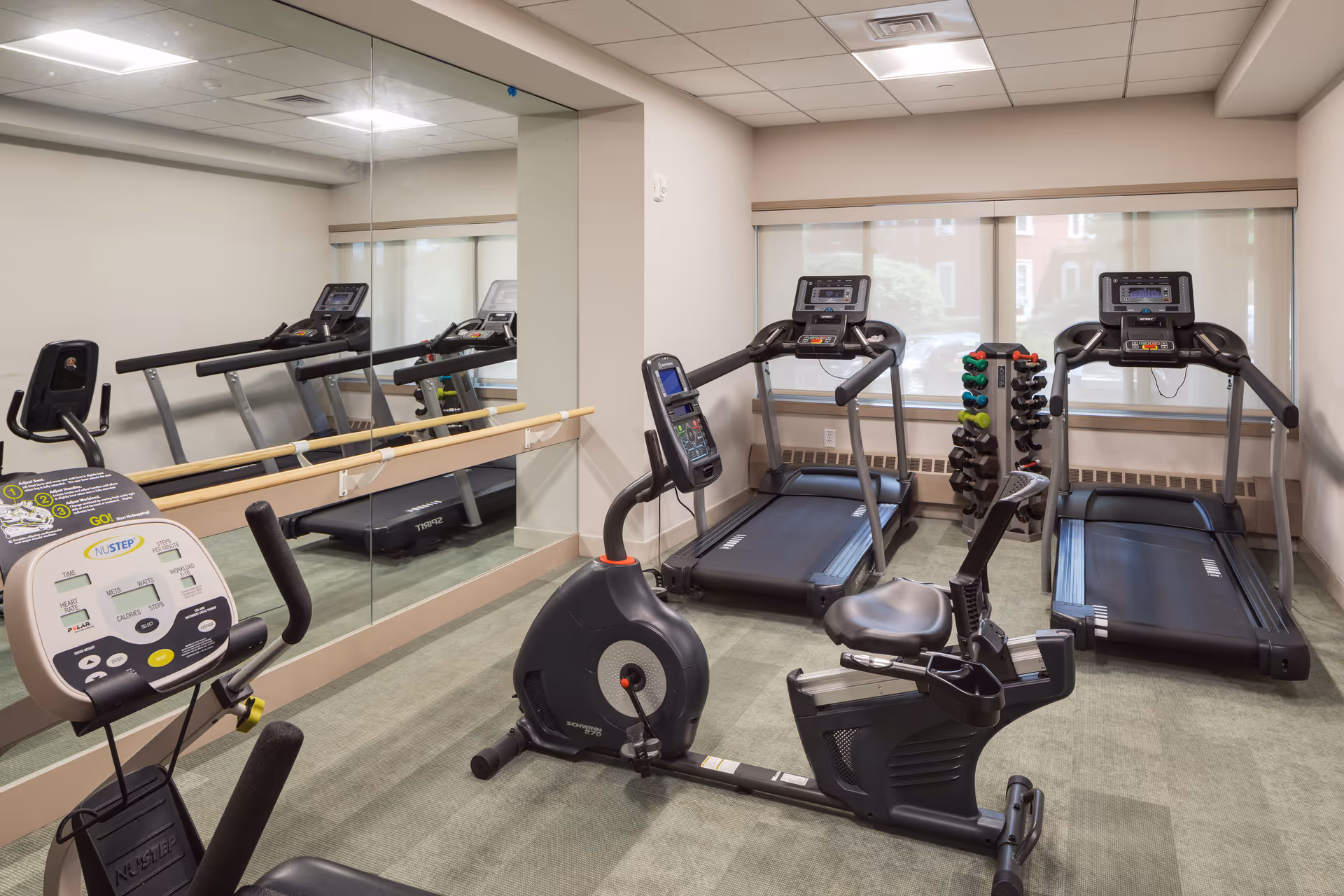 A small fitness room with two treadmills, a recumbent exercise bike, a NuStep machine, a rack of various dumbbells, and a large wall mirror reflecting the equipment. The room has beige walls, carpeted floor, and windows with blinds partially drawn.