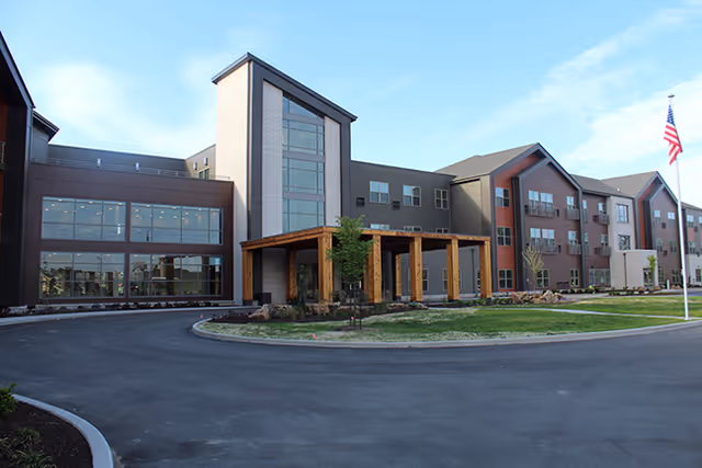 Exterior view of a modern three-story senior living facility with large windows, a covered entrance supported by wooden beams, landscaped grounds, and an American flag on a flagpole in front.