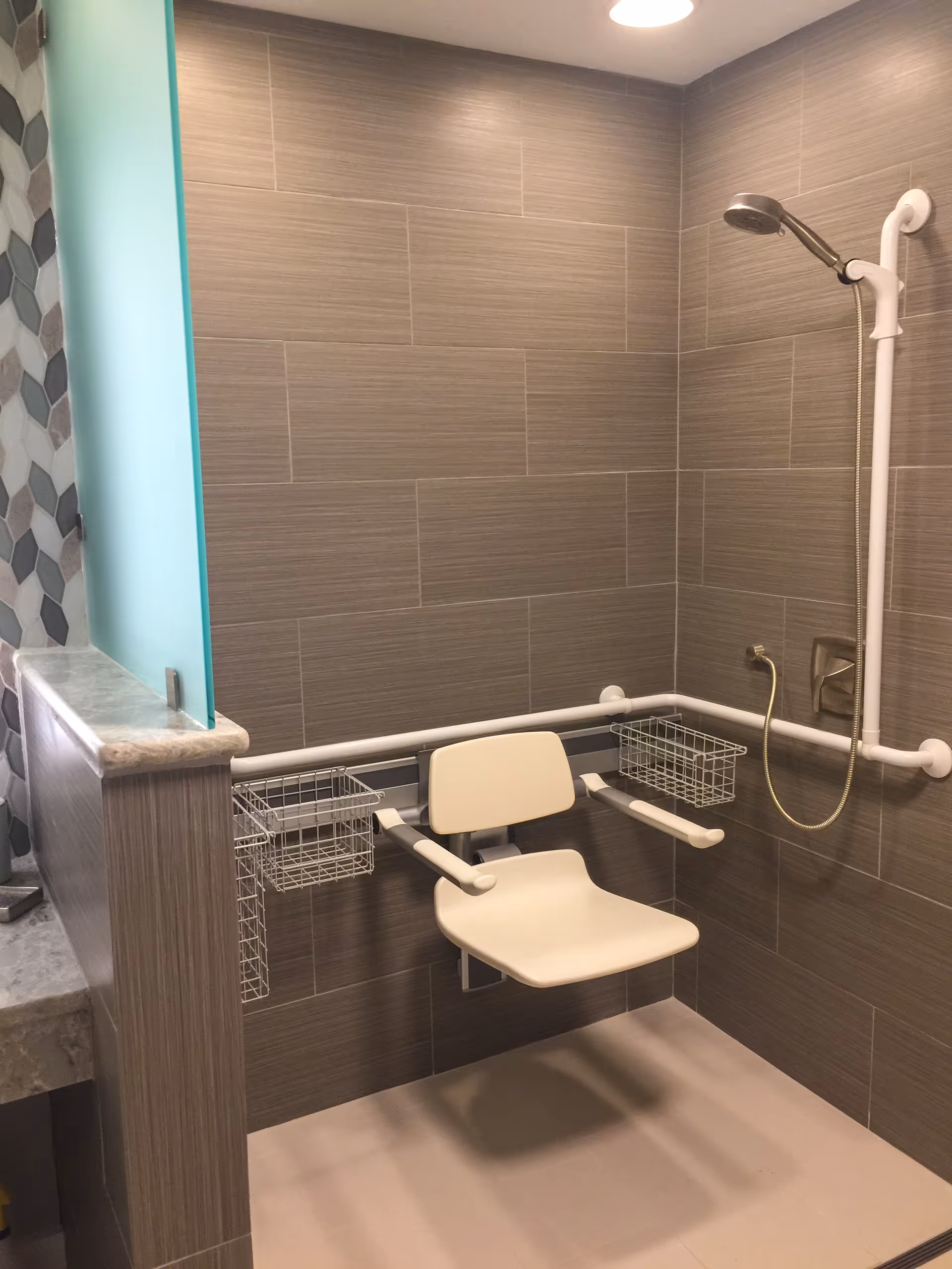 A modern shower area featuring a wall-mounted showerhead, a fold-down seat, and grab bars for support, with a sleek tile design.