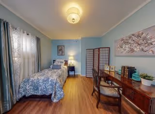 A cozy bedroom with a single bed covered in blue and white patterned bedding, a nightstand with a lamp, a wooden desk with a chair, decorative items including framed pictures and plants, light blue walls, wood flooring, and a window with sheer curtains.