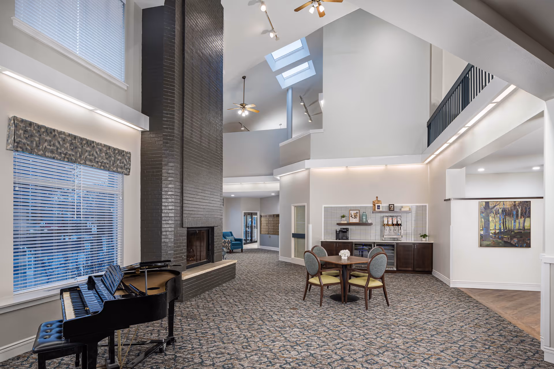 Spacious senior living facility common area with a grand piano, a tall black brick fireplace, a round table with four chairs, and a beverage station against the wall. The room features high ceilings with skylights, ceiling fans, large windows with blinds, and patterned carpet flooring.