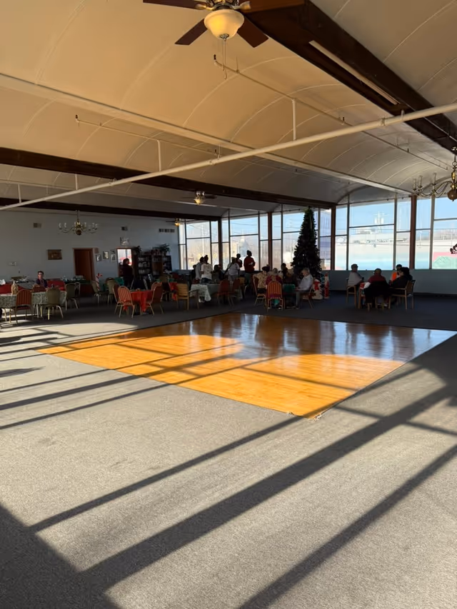 A large room with a wooden dance floor in the center and carpeted area surrounding it. Several tables with chairs are arranged around the room, with people sitting and socializing near the windows. A decorated Christmas tree is visible near the windows, and the room has large windows letting in natural light. Ceiling fans and chandeliers hang from the ceiling.