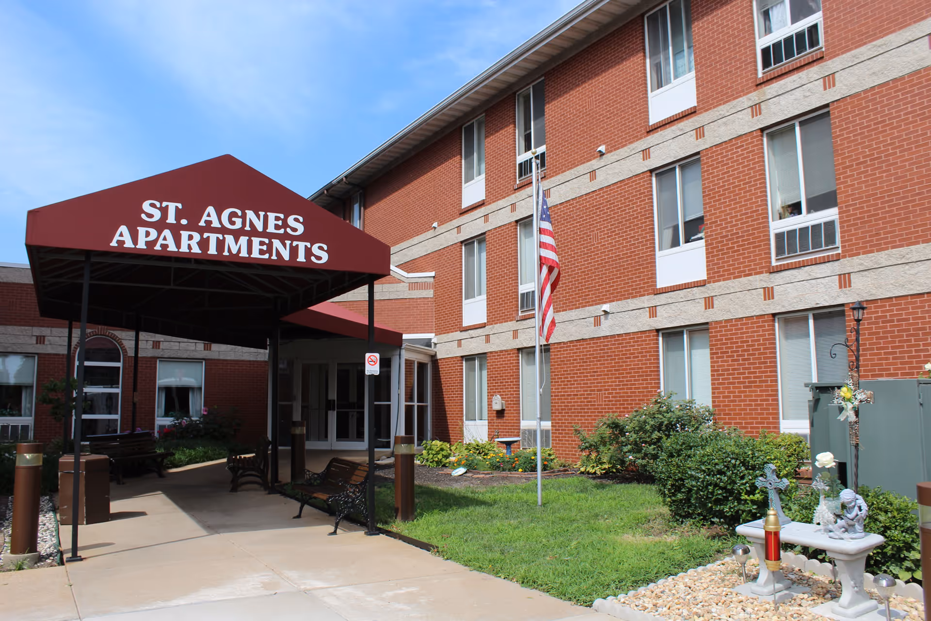 Exterior view of St. Agnes Apartments showing a red brick building with multiple windows, a covered entrance with a maroon canopy displaying the name 'St. Agnes Apartments', an American flag on a flagpole, and a small garden area with bushes and decorative items.