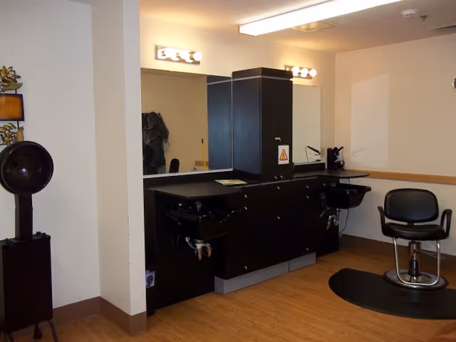 Interior view of a hair salon area within a senior living facility, featuring two black salon chairs, two black hair washing sinks, a large mirror with lights above, and wooden flooring.